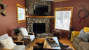 A cozy living room with a stone fireplace, a mounted TV, and a large window with blinds.