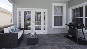 Un patio con una parrilla negra, un sofá con cojines estampados y una puerta de vidrio que da a una zona de piscina.