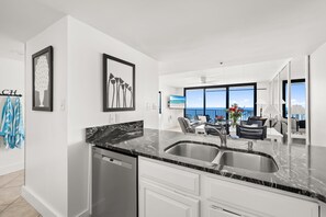 A modern kitchen with a black marble countertop, stainless steel sink, and white cabinetry. There is a dining area with a view of the ocean through large windows.