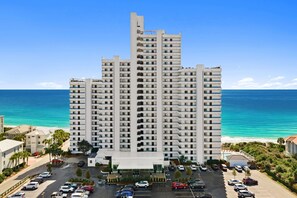 A multi-story hotel with a central entrance, parking lot, and ocean view.