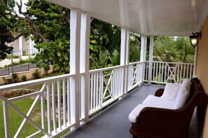 A covered porch with white railings, a wicker sofa with cushions, and a view of lush greenery and a building in the background.
