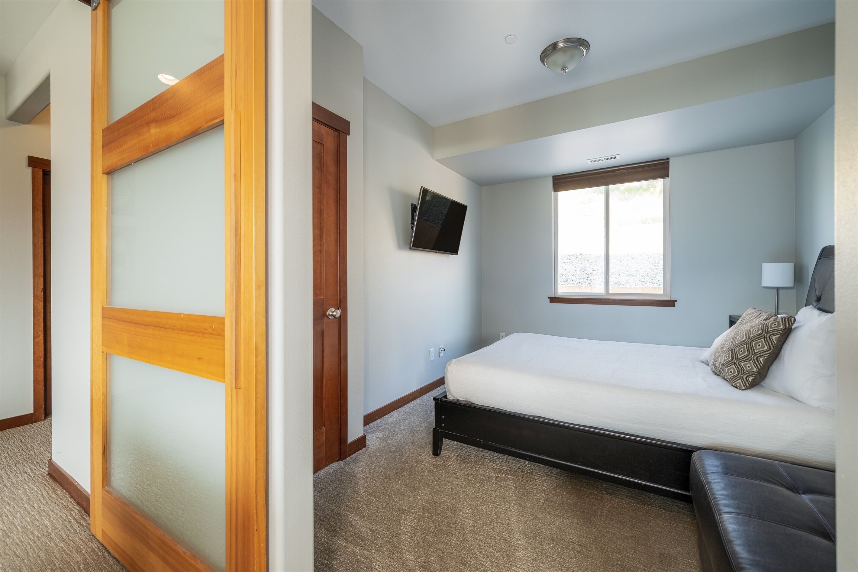 Un dormitorio con cama, televisor montado en la pared, ventana y puerta.
