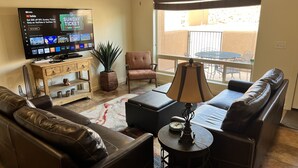 A living room with a leather sofa, a coffee table, a TV displaying NFL Sunday Ticket, a potted plant, and a view of a balcony with outdoor furniture. - 8A6 Amazing Views, Shared Pool/Hot Tub, Private Patio and Garage (MOAB)