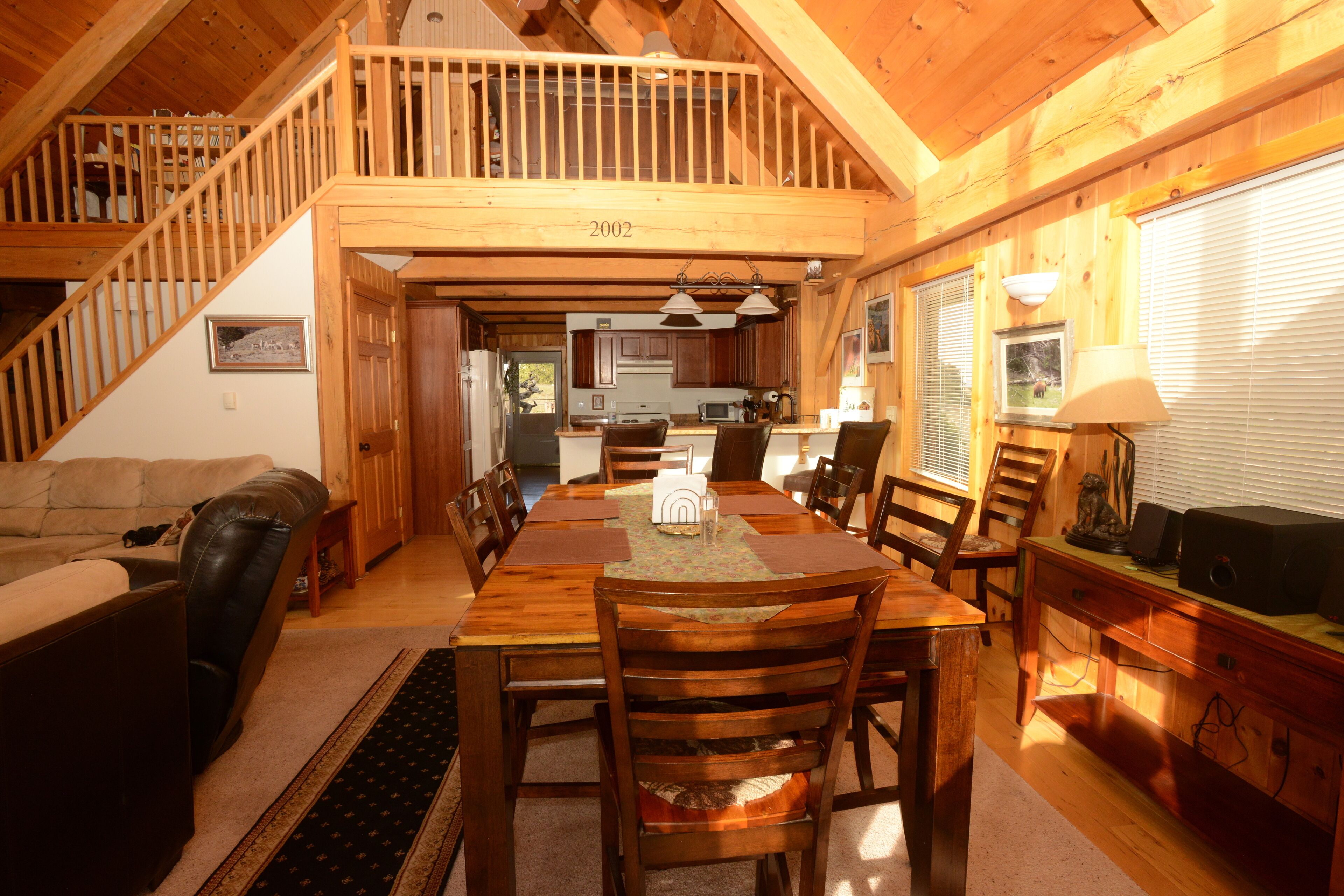 A spacious dining area with a high ceiling and wooden beams, a kitchen in the background, and a staircase leading to an upper level.