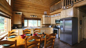 A wooden cabin kitchen with a dining table set for four, a refrigerator, and wooden cabinets. - Cpt Cook - Unique Alaskan log home in trees & wildflowers on 14-acre homestead. (Anchor Point)