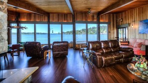 Amplia sala de estar con sofás de cuero, comedor y grandes ventanales con vista a un lago.