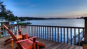 Una terraza de madera con vista a un lago tranquilo, rodeada de árboles y una orilla lejana.