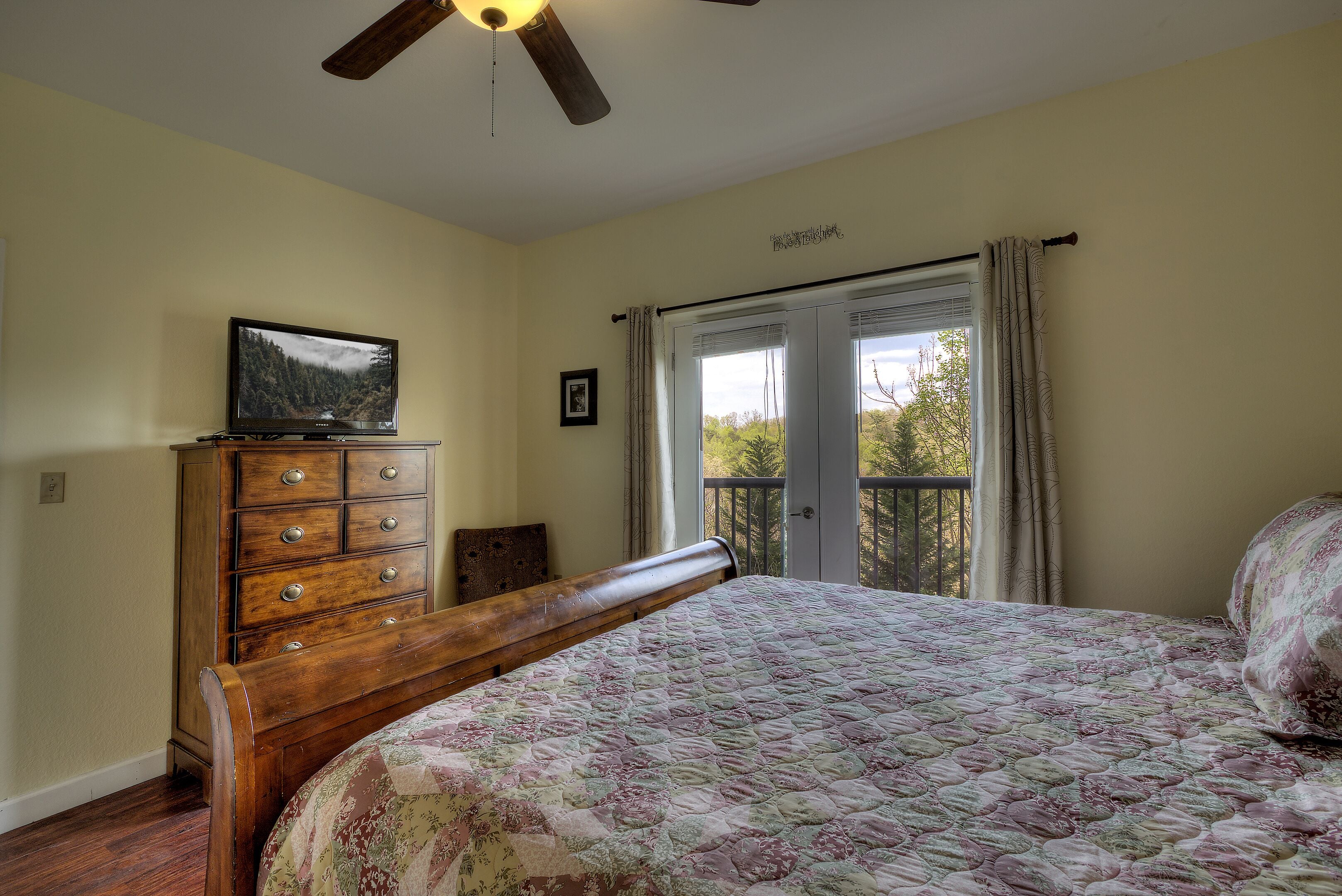 A bedroom with a wooden bed, a dresser, a television on top, a ceiling fan, and a window with curtains.