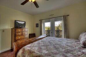 Une chambre avec un lit en bois, une commode, un téléviseur posé dessus, un ventilateur de plafond et une fenêtre avec des rideaux.