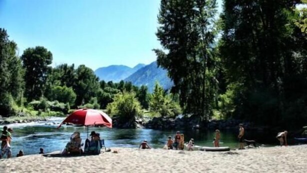 A river with people relaxing on the sand, a red umbrella, and trees in the background.