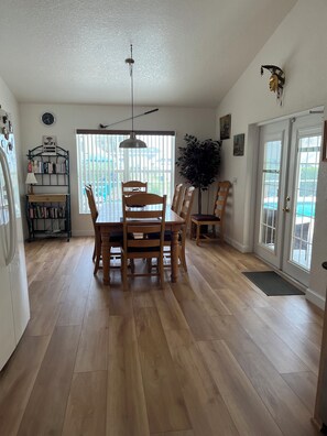 A dining area with a wooden table and chairs, a hanging light fixture, a bookshelf, and a large window with blinds.