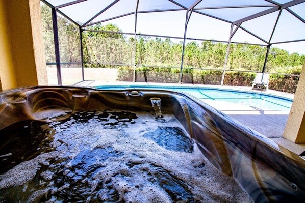 A hot tub with a transparent roof, surrounded by a pool and a view of a forested area.
