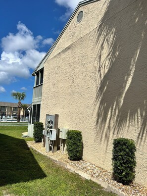 A building with a textured facade, a small window, and a door. There are two bushes and a small utility box in front.