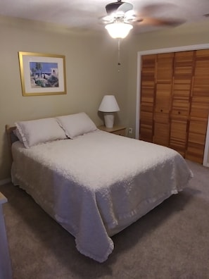 A bedroom with a bed, a nightstand, a lamp, a ceiling fan, a wooden wardrobe, and a framed picture on the wall.