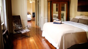 A bedroom with a wooden floor, a large bed with white bedding, a wooden wardrobe, and a chair.