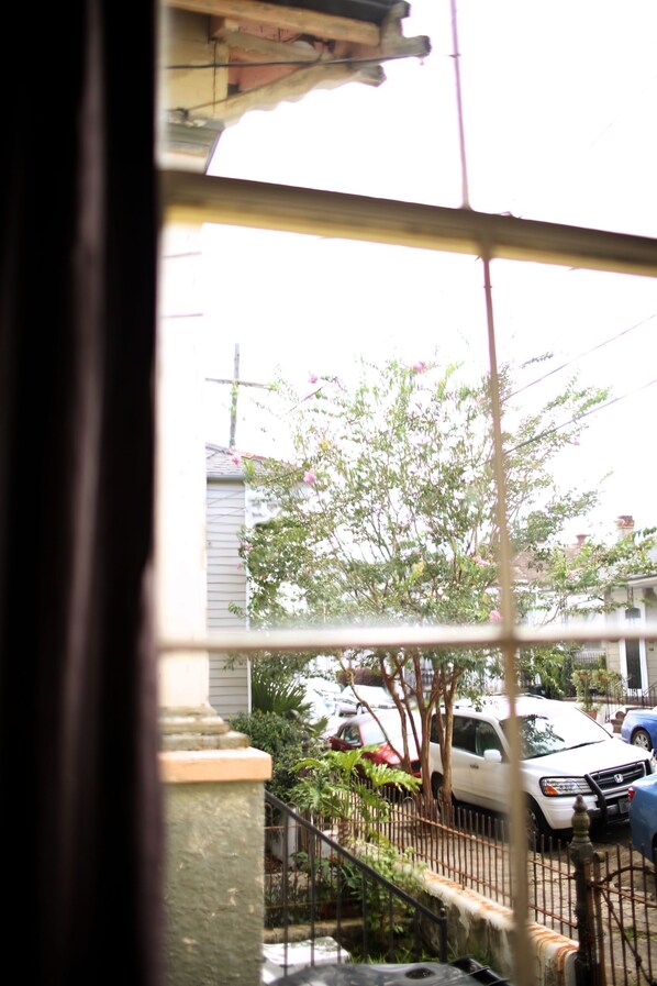 Vista a través de una ventana de metal, mostrando un coche blanco estacionado en una entrada de casa, un árbol con flores rosadas y una cerca metálica.