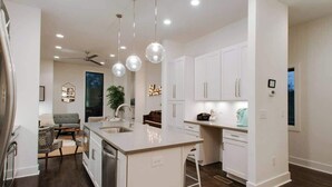 Una cucina moderna con mobili bianchi, un'isola centrale, illuminazione a sospensione e un'area pranzo con divano grigio e sedie a fantasia.
