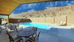 Zona de piscina al aire libre con un porche cubierto, mesas y sillas.