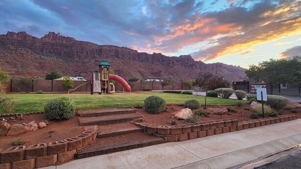 Un parque infantil con tobogán y estructura de escalada, rodeado de un jardín ajardinado con escalones y rocas, con montañas y un cielo al atardecer como telón de fondo.