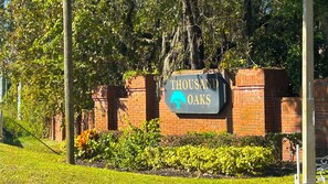 A sign with 'THOUSAND OAKS' and a tree silhouette, surrounded by brick walls and greenery. - HUGE Pool Deck 15 mins to Disney! 360º Sun, Privacy-Games-Chef’s Kitchen + More! (Loughman)