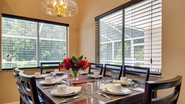A dining area with a table set for four, two chairs on each side, a vase with flowers, and large windows with blinds.