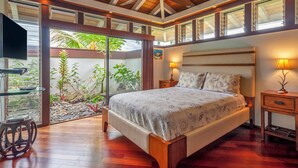 Un dormitorio con una cama grande, suelo de madera, ventilador de techo y vistas a un jardín a través de puertas de cristal.