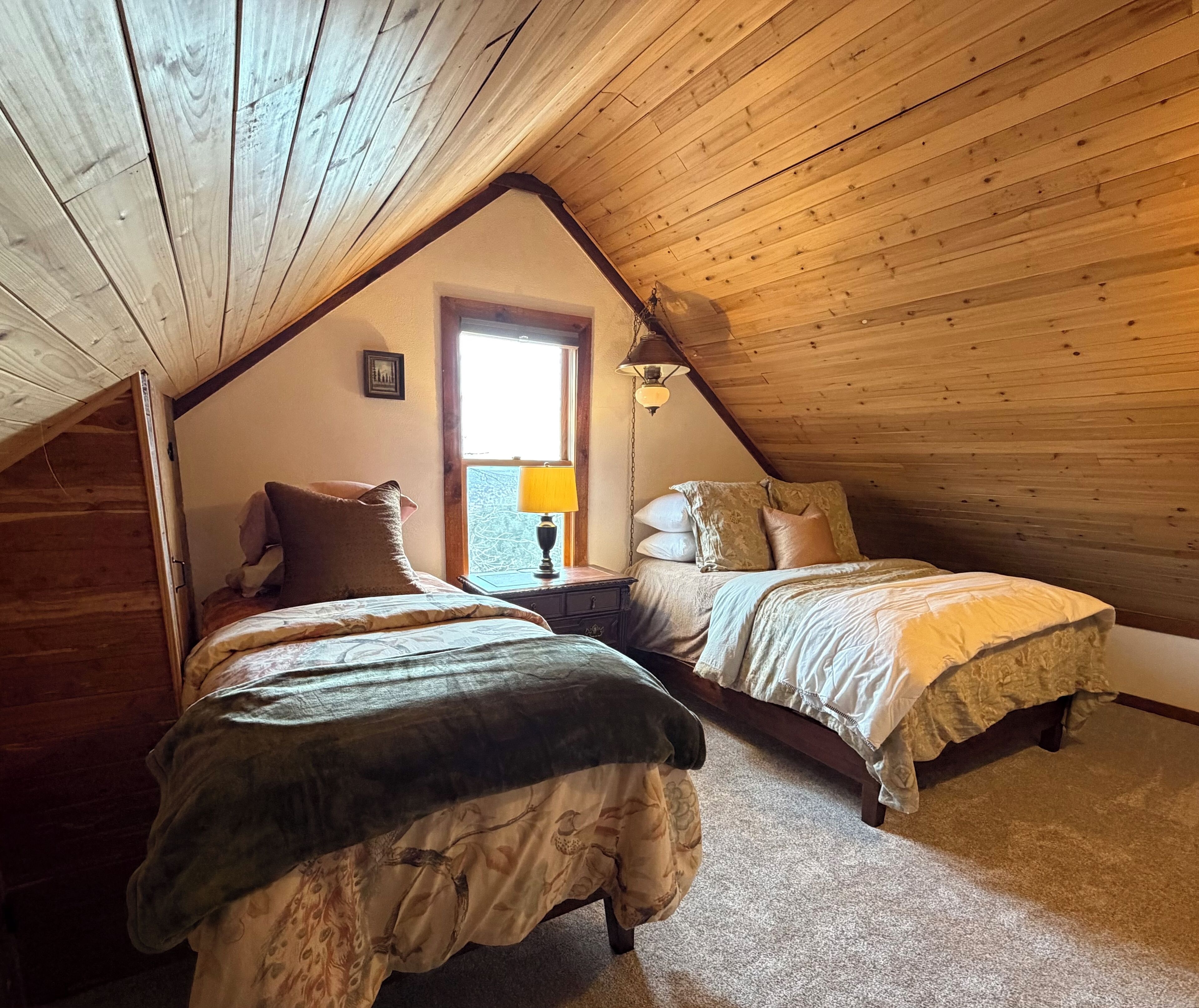 A cozy attic bedroom with two beds, wooden ceiling beams, a window with a view, and a lamp on a nightstand.