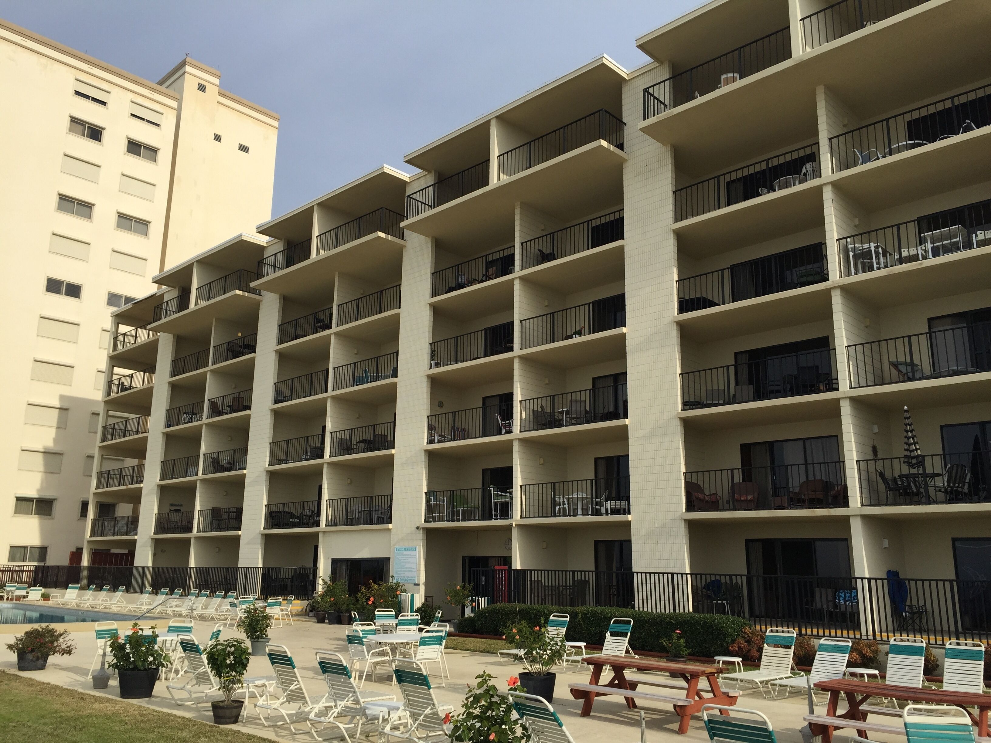 A multi-story residential building with balconies, a pool area with lounge chairs, and potted plants.