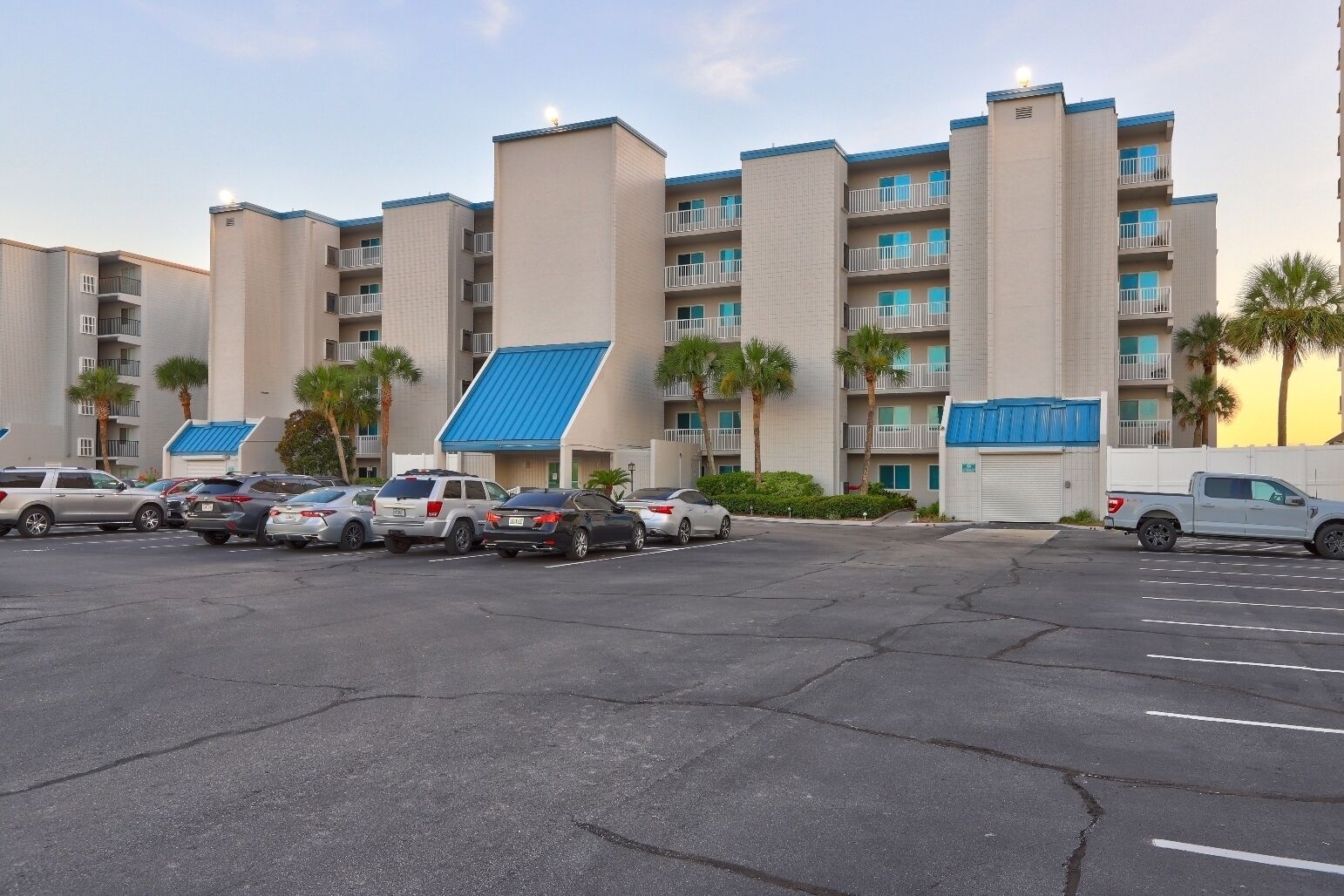 A parking lot with several cars and a building with multiple balconies and a blue roof.