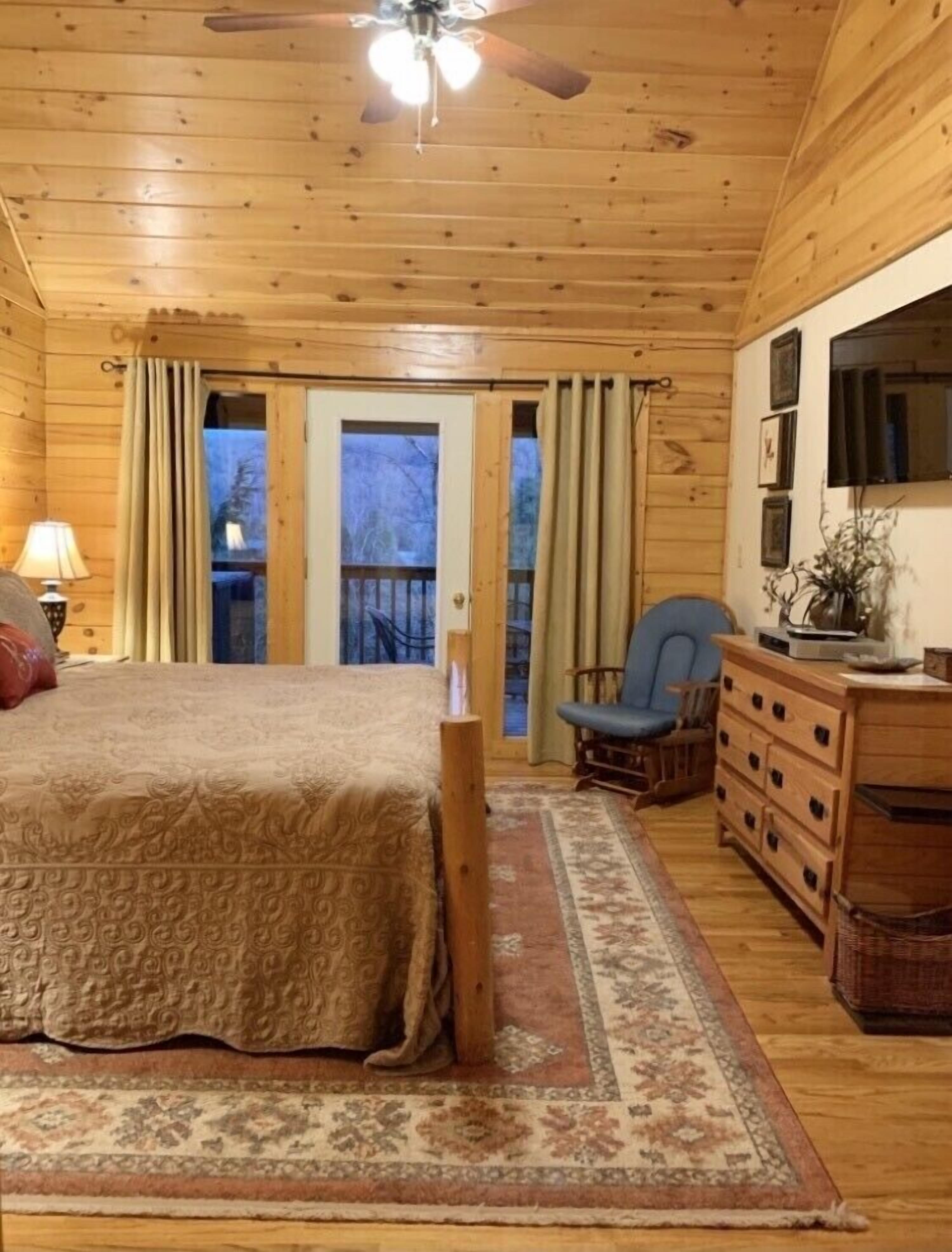 A wooden cabin bedroom with a bed, dresser, chair, and a ceiling fan.