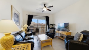 A living room with a black leather sofa, a glass coffee table, a wooden side table, a TV on a wooden stand, and a ceiling fan. - Tinkerbell's Hideaway 2.5 miles from Disney/pool/NO resort fees/FREE parking (Kissimmee)