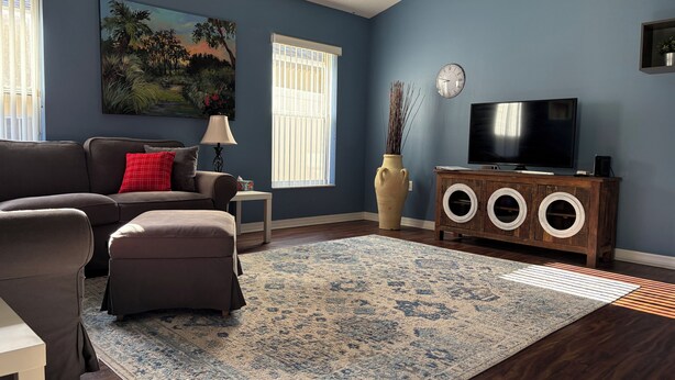 Una sala de estar con sofá, reposapiés, alfombra, mueble para televisión y un cuadro en la pared.