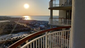 A balcony with a view of the sunset over a body of water, a parking area, and a residential area below. - PENTHOUSE 17th floor w. free Beach Service (chairs/umbrella) included! (Navarre)