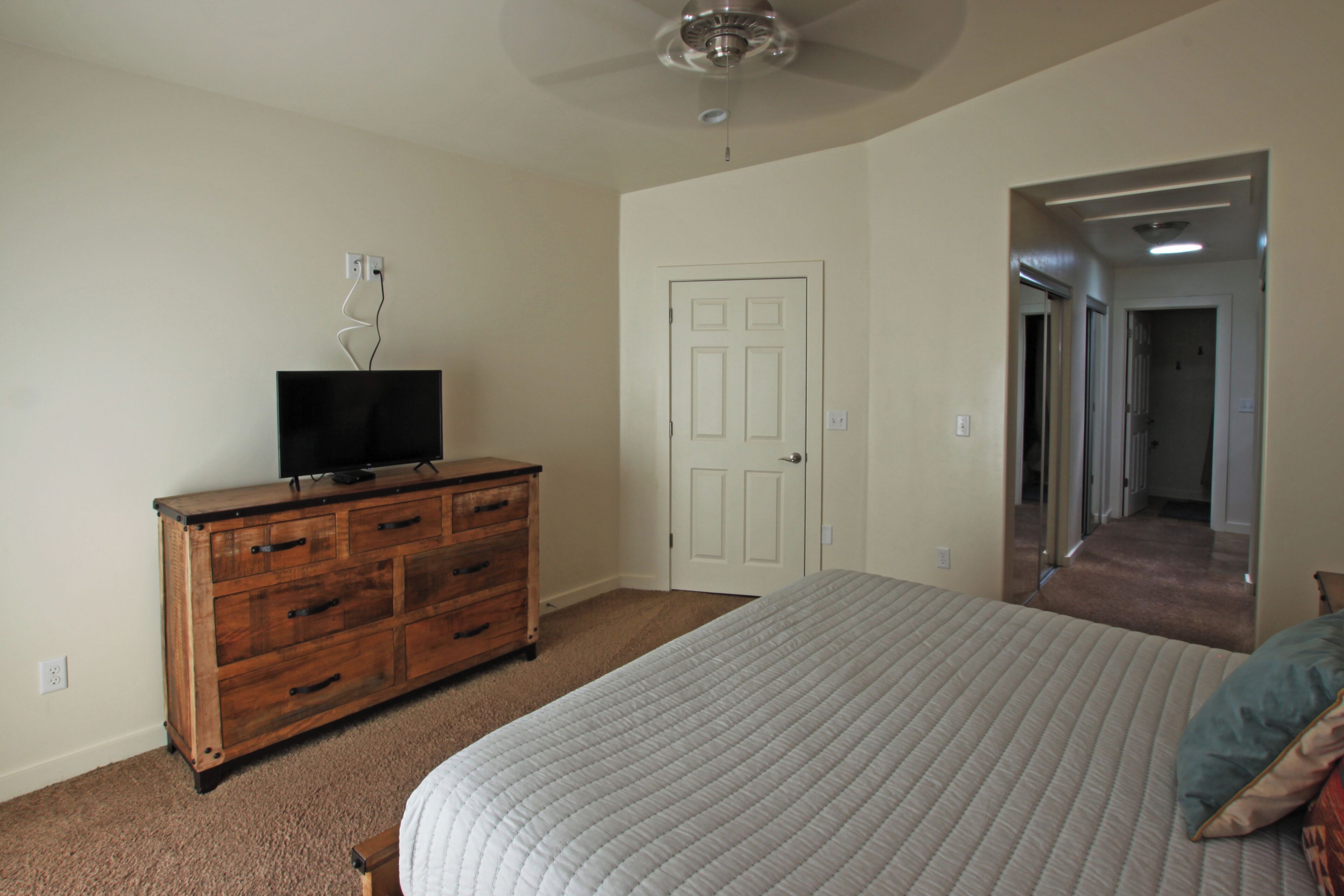 A bedroom with a large bed, a wooden dresser, a television on top, and a door leading to another room.