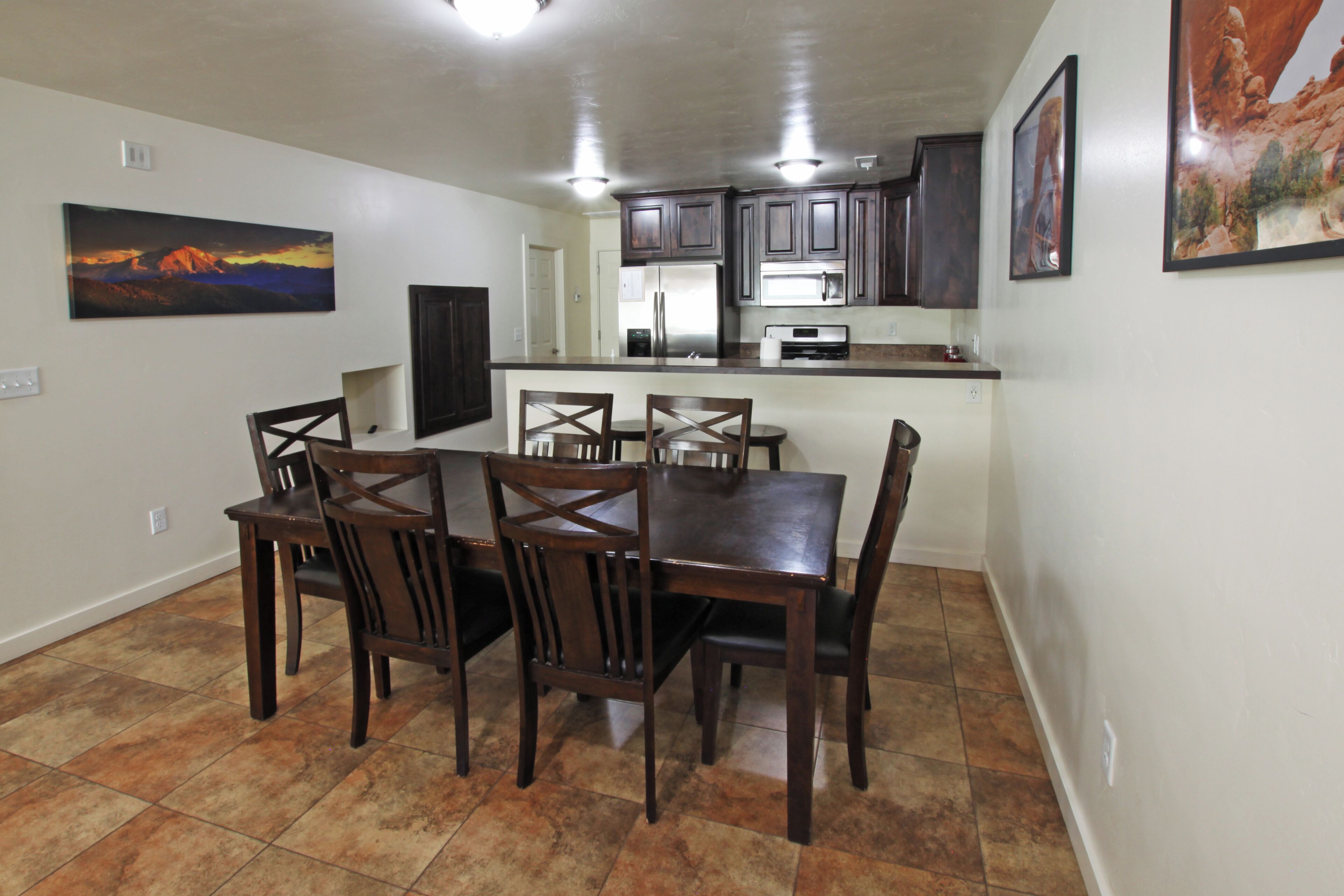 A dining area with a large wooden table and chairs, a kitchen with dark cabinets and stainless steel appliances, and artwork on the walls.