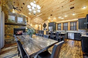 Un vaste espace dédié à la salle à manger, avec une grande table et des chaises en bois, une cheminée en pierre et une cuisine dotée de meubles sombres et de grandes fenêtres.