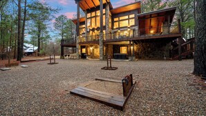 A multi-story log cabin with a stone foundation, a balcony, and a gravel driveway. - 4 Bedroom 3.5 Bath-Pickleball/Basketball Court, Game Room, Hot Tub & Playground (Broken Bow)