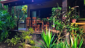 A covered outdoor seating area with wooden chairs and tables, surrounded by lush greenery and potted plants.