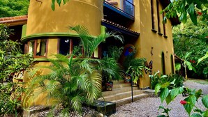 A yellow building with a balcony, surrounded by lush greenery and potted plants.