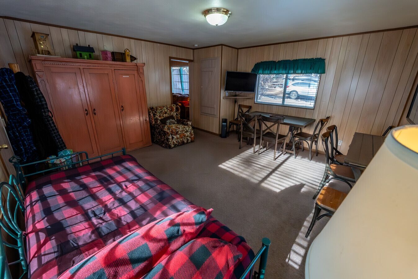 A room with a bed, a TV, a dining table with chairs, a wardrobe, and a window with a view of a parked car.