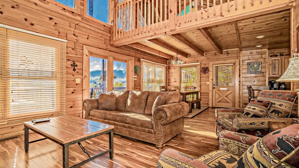A log cabin interior with a wooden floor, a brown leather sofa, a matching armchair, a wooden table, and a balcony with a railing. - 5 miles to Gatlinburg, Hot Tub, Views, 4 BEDROMS: JEEP INVASION, ROD RUN, SPARKS (Pigeon Forge)