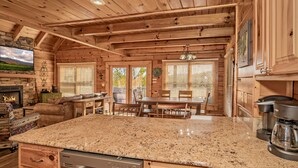A wooden cabin interior with a kitchen area featuring granite countertops, a dining table, and a fireplace. - 5 miles to Gatlinburg, Hot Tub, Views, 4 BEDROMS: JEEP INVASION, ROD RUN, SPARKS (Pigeon Forge)