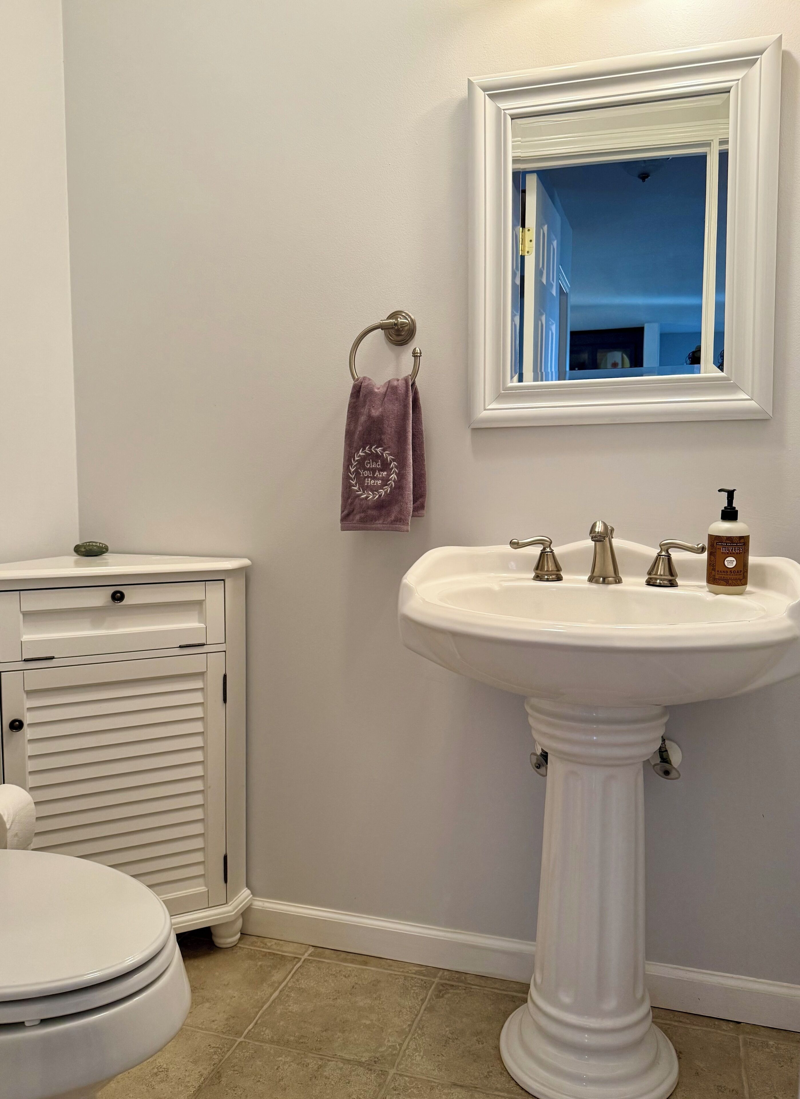 Un baño con un lavamanos pedestal blanco, un inodoro, un mueble con cajones, una toalla colgada en un toquillero y un espejo sobre el lavamanos.