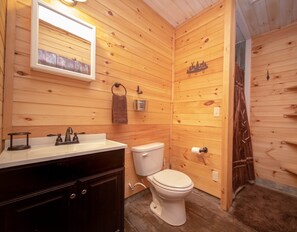 Un baño con paneles de madera, un lavamanos, un espejo, un perchero para toallas, un inodoro y una puerta de madera.