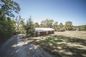 Una cabaña rústica con camino de grava, rodeada de árboles y un campo cubierto de hierba.