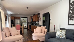 Una sala de estar con un sofá gris oscuro, dos sillones beige y una mesa de centro de madera. Se ve una cocina al fondo, con armarios de madera y un refrigerador.