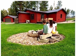 A red wooden cabin with a stone fire pit in front.