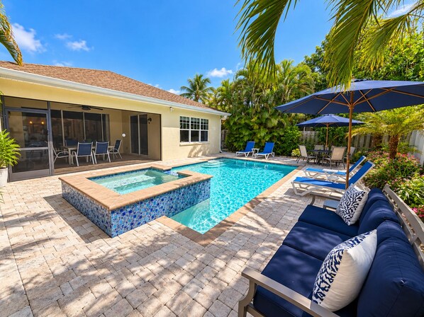 A backyard with a swimming pool, a hot tub, and a patio area with lounge chairs and umbrellas.