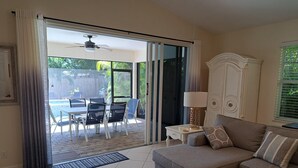 A living room with a grey sofa, a coffee table, and a dining area with a table and chairs. There is a sliding glass door leading to a patio with a pool and a ceiling fan. - Naples Best Value Luxury 4 Bedroom Heated Pool Spa Bikes Spacious Backyard (Naples)