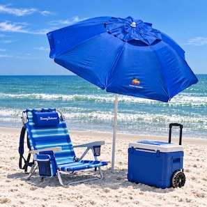 A beach chair with a blue umbrella, a cooler, and a beach setting.